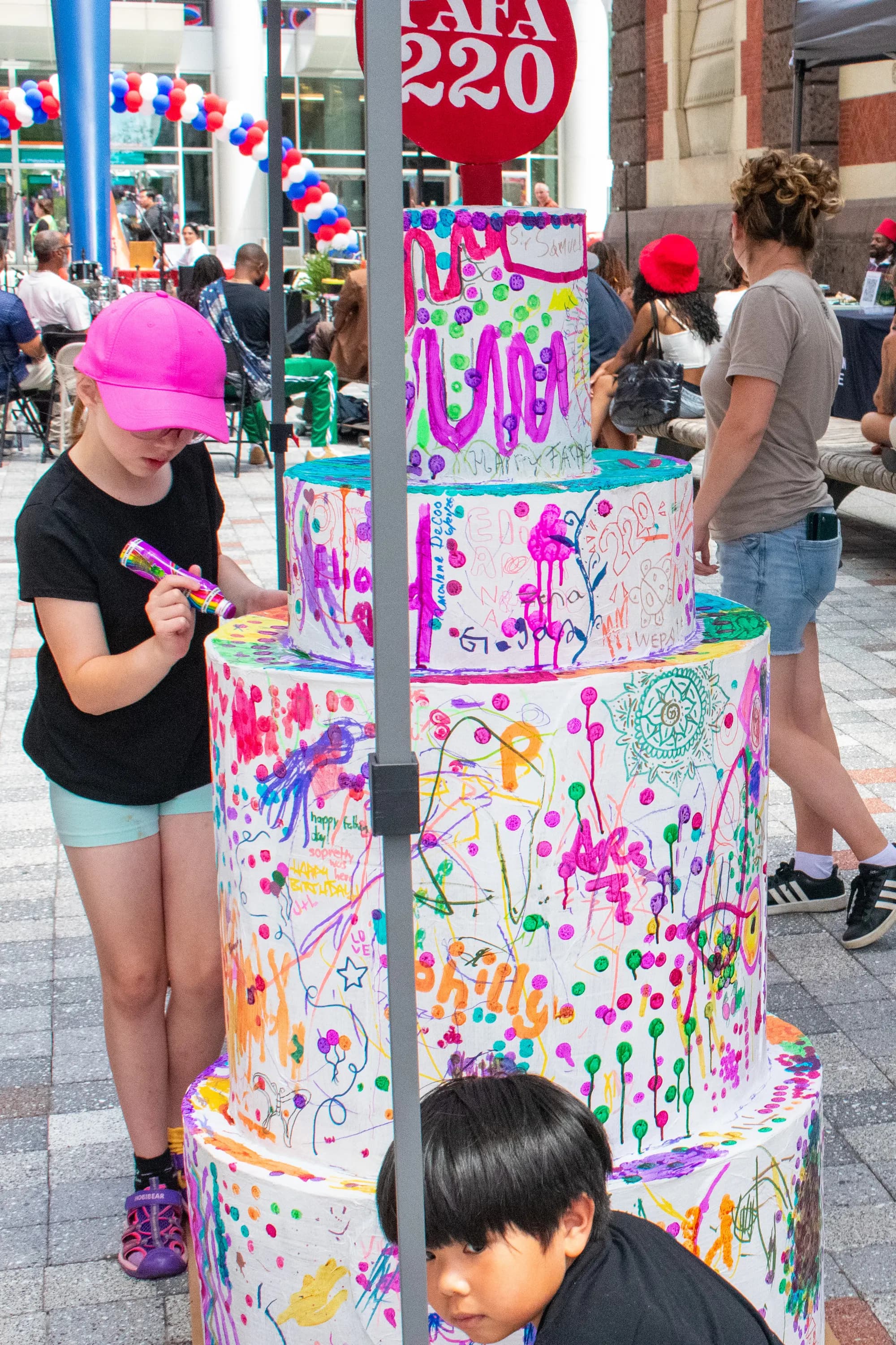 A child in a pink bucket hat painting a large sculpted cake already covered in bright splatters.