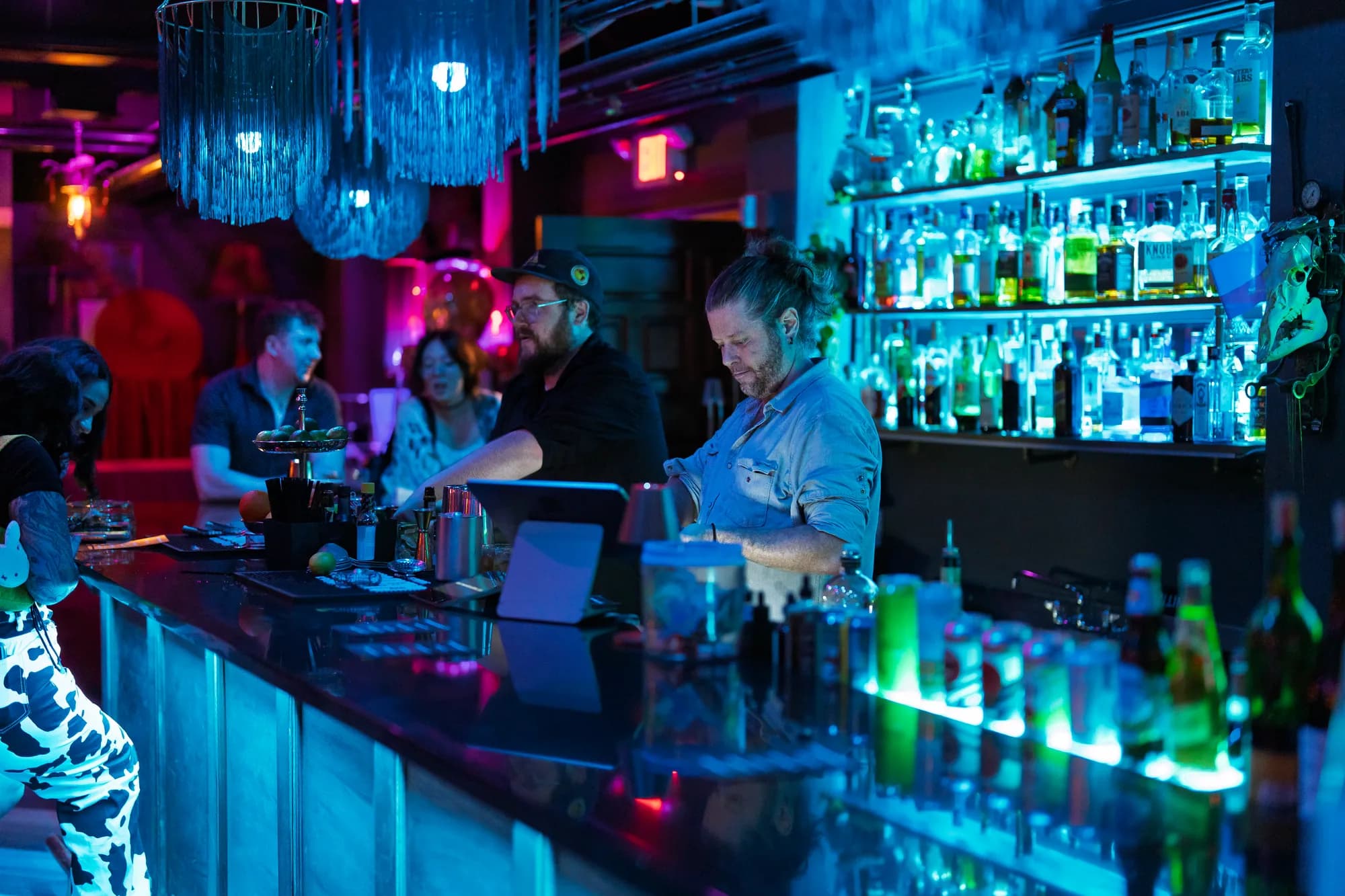 Bartenders at work behind the long bar, back shelves lit in blue under fringed chandeliers.
