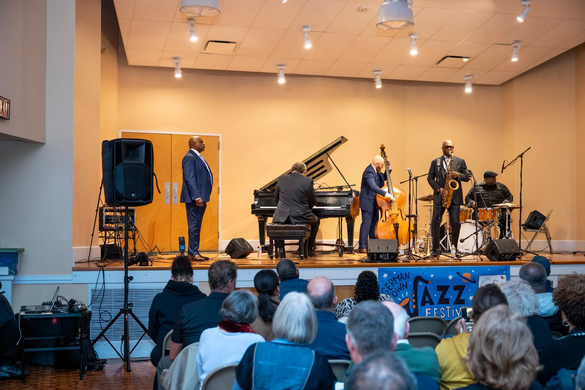 A trio on stage — piano, upright bass, and saxophone — playing to a seated audience.