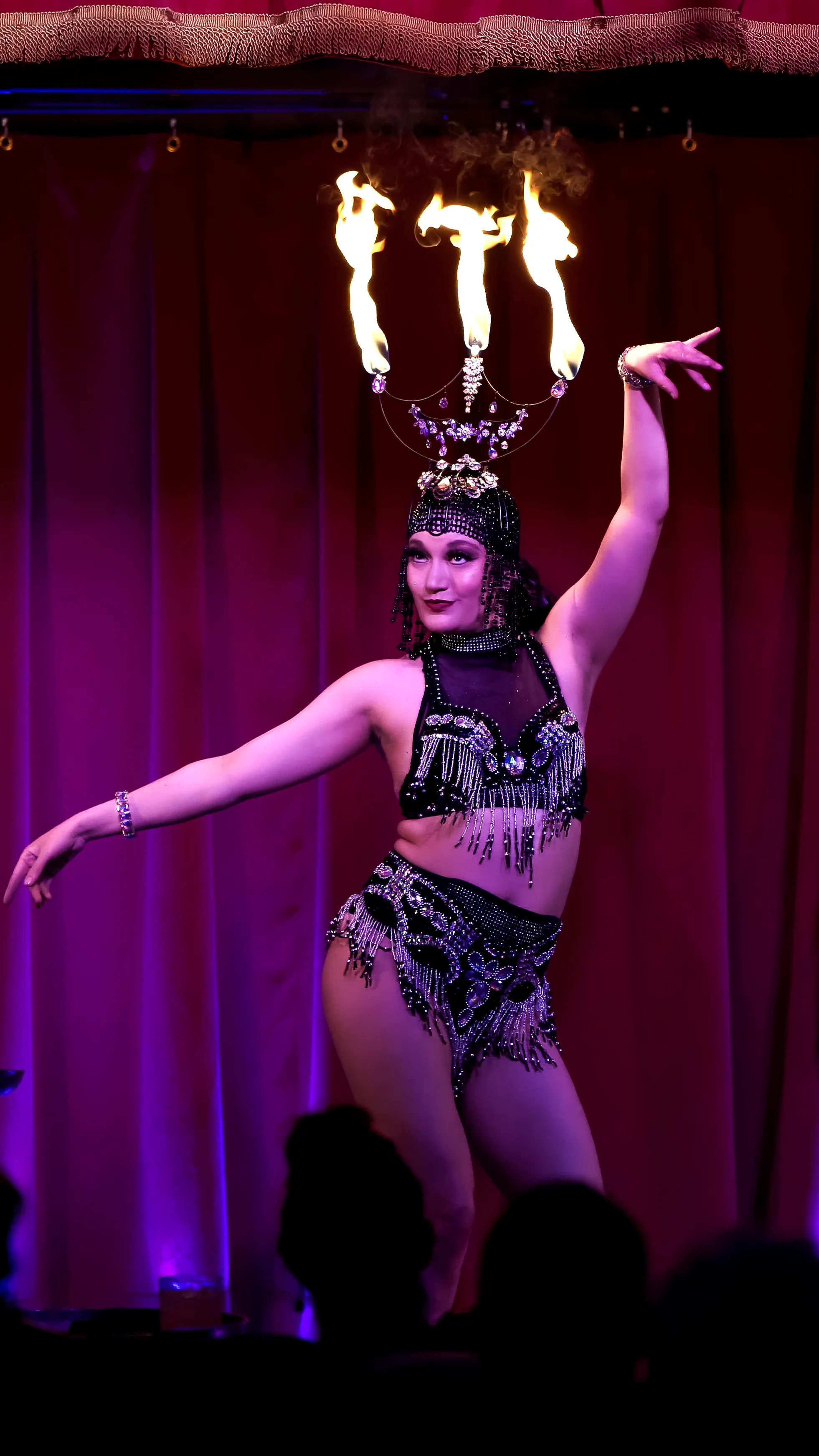 A burlesque dancer arching under a headdress of live flames on the red-curtained stage.
