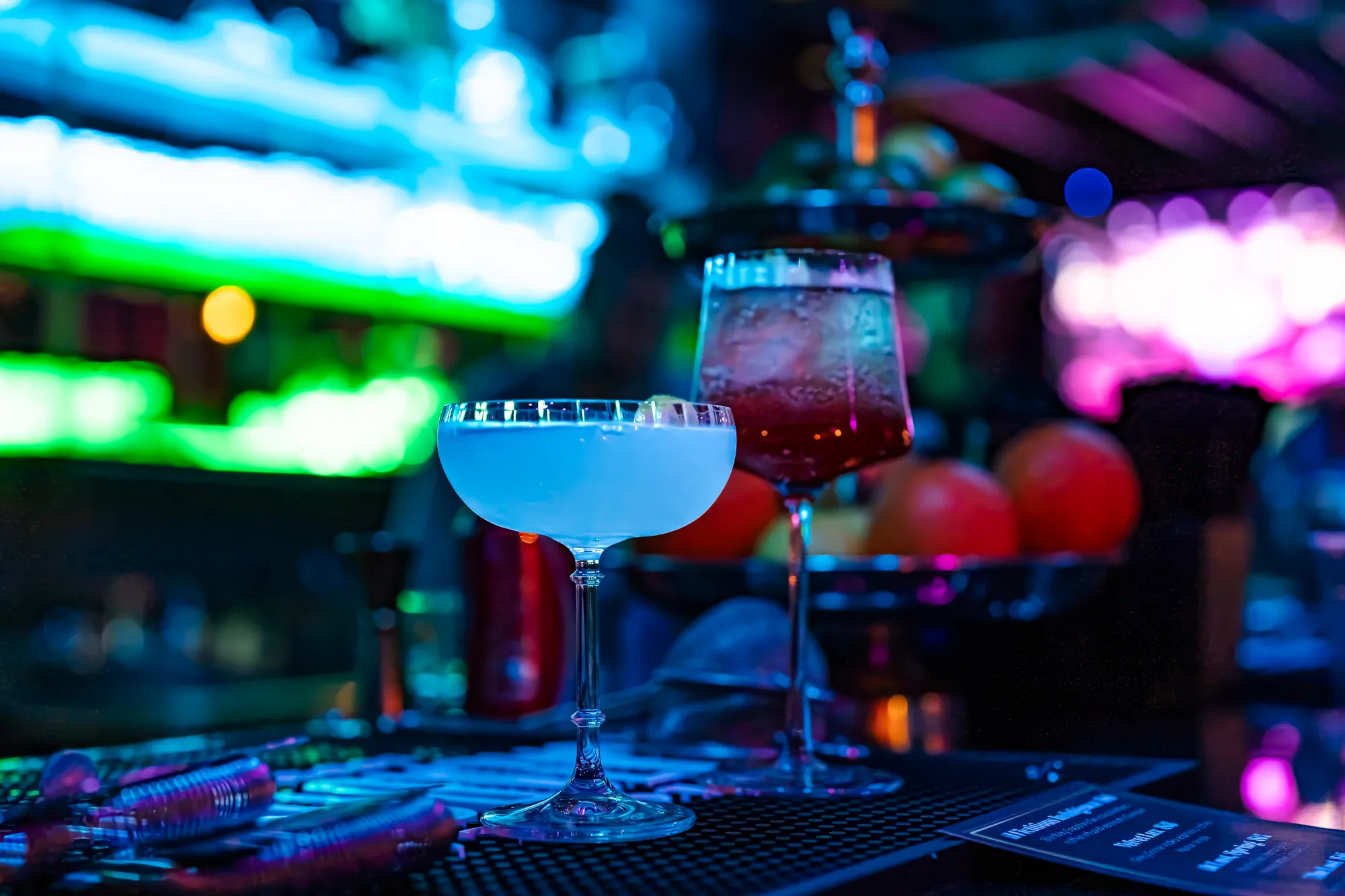 Two cocktails on the bar top, a pale coupe and a red spritz, neon blurring behind them.