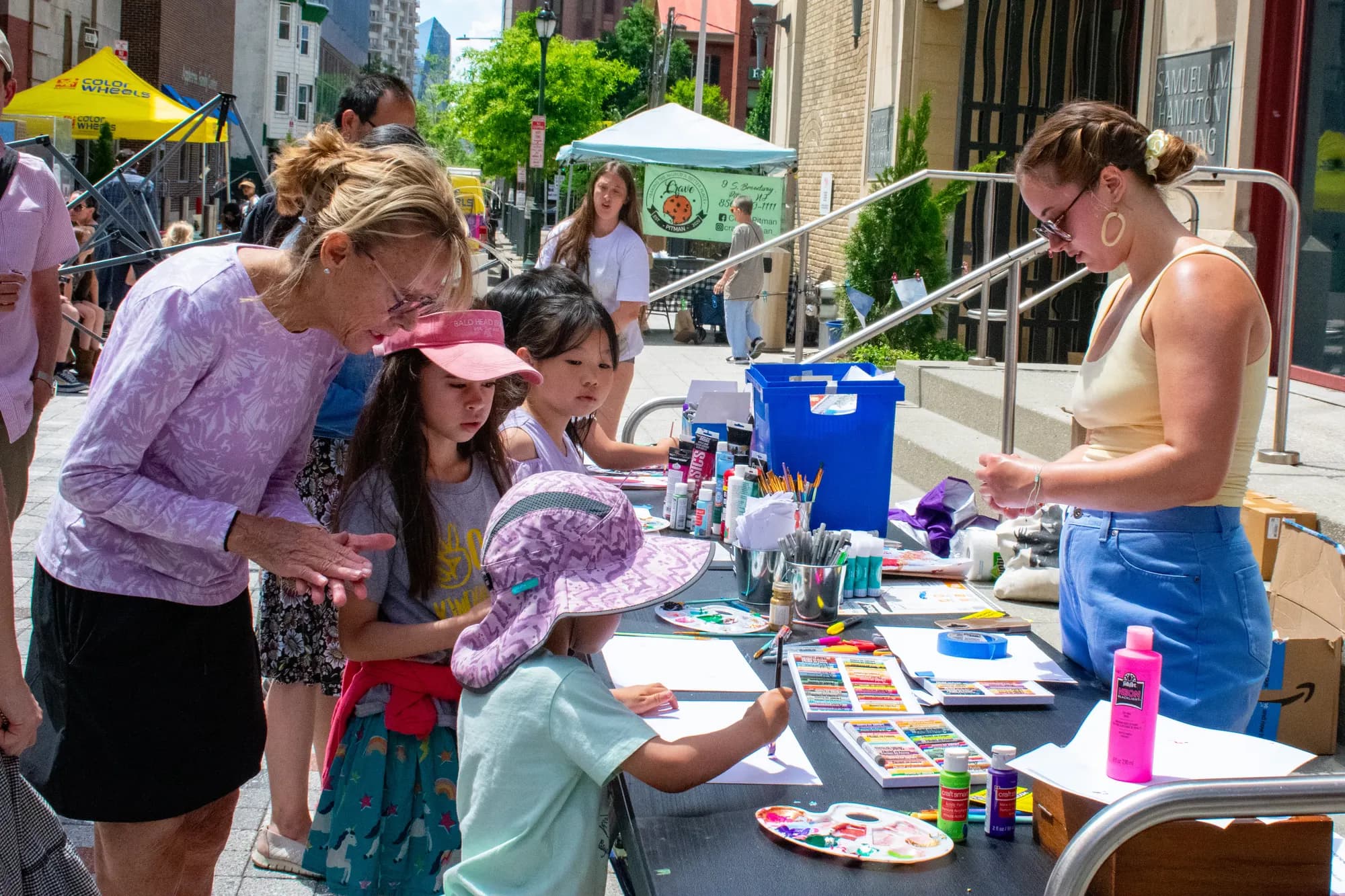 Adults and children painting side by side at an outdoor table set with watercolor palettes.