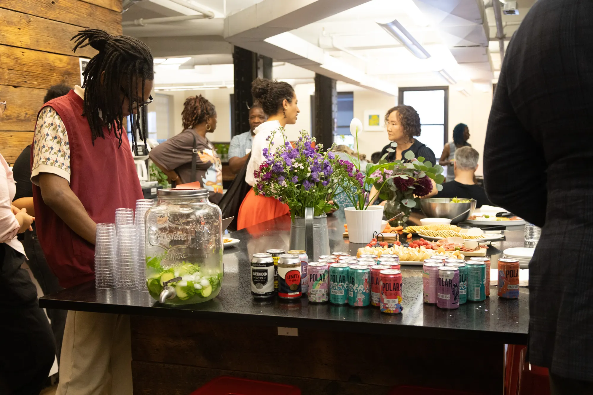 The refreshment table set with an infused-water dispenser, ranks of canned drinks, and cut flowers, guests talking behind it.