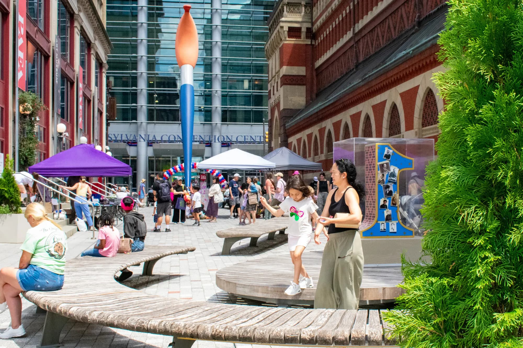A wide view of the plaza, the paintbrush sculpture rising over the festival tents.