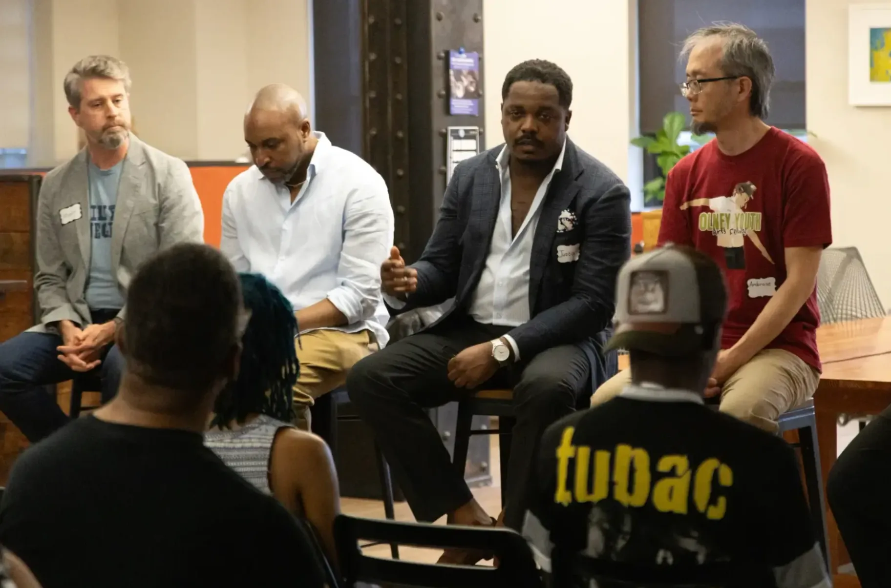 Four panelists seated before the audience, the speaker in a navy jacket gesturing as he makes his point.