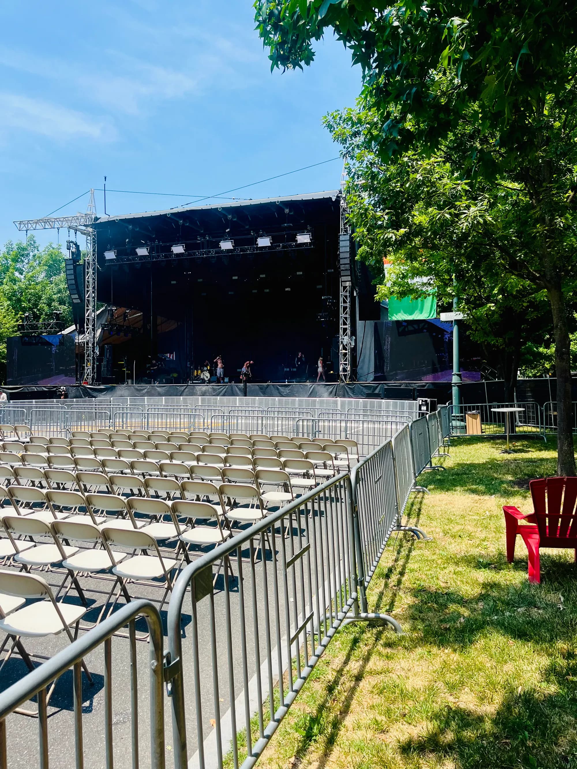 Rows of white folding chairs behind crowd barriers facing the main stage, a band soundchecking under a clear July sky.