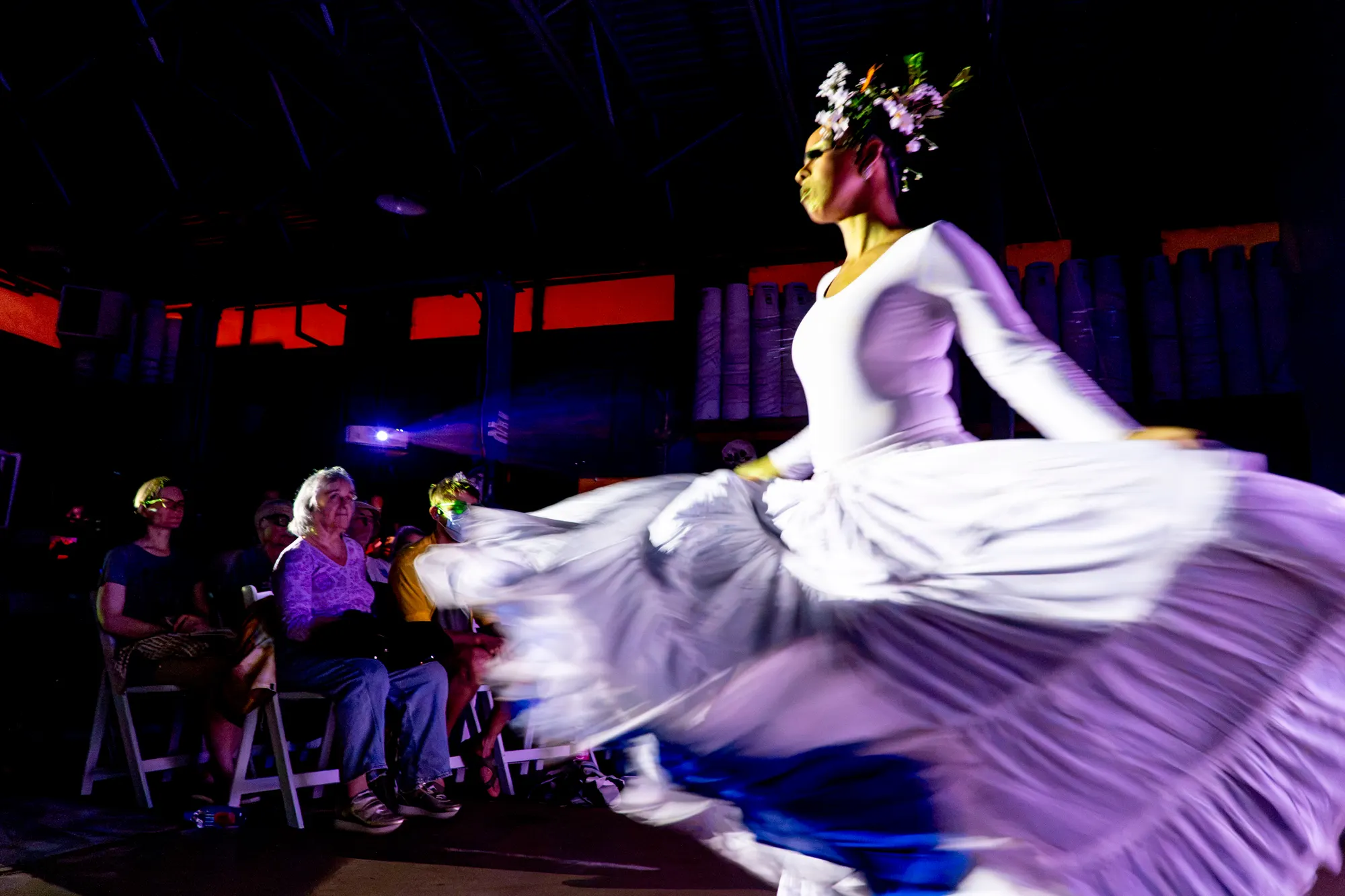 A dancer in a white and violet dress turning mid-phrase on a darkened stage.