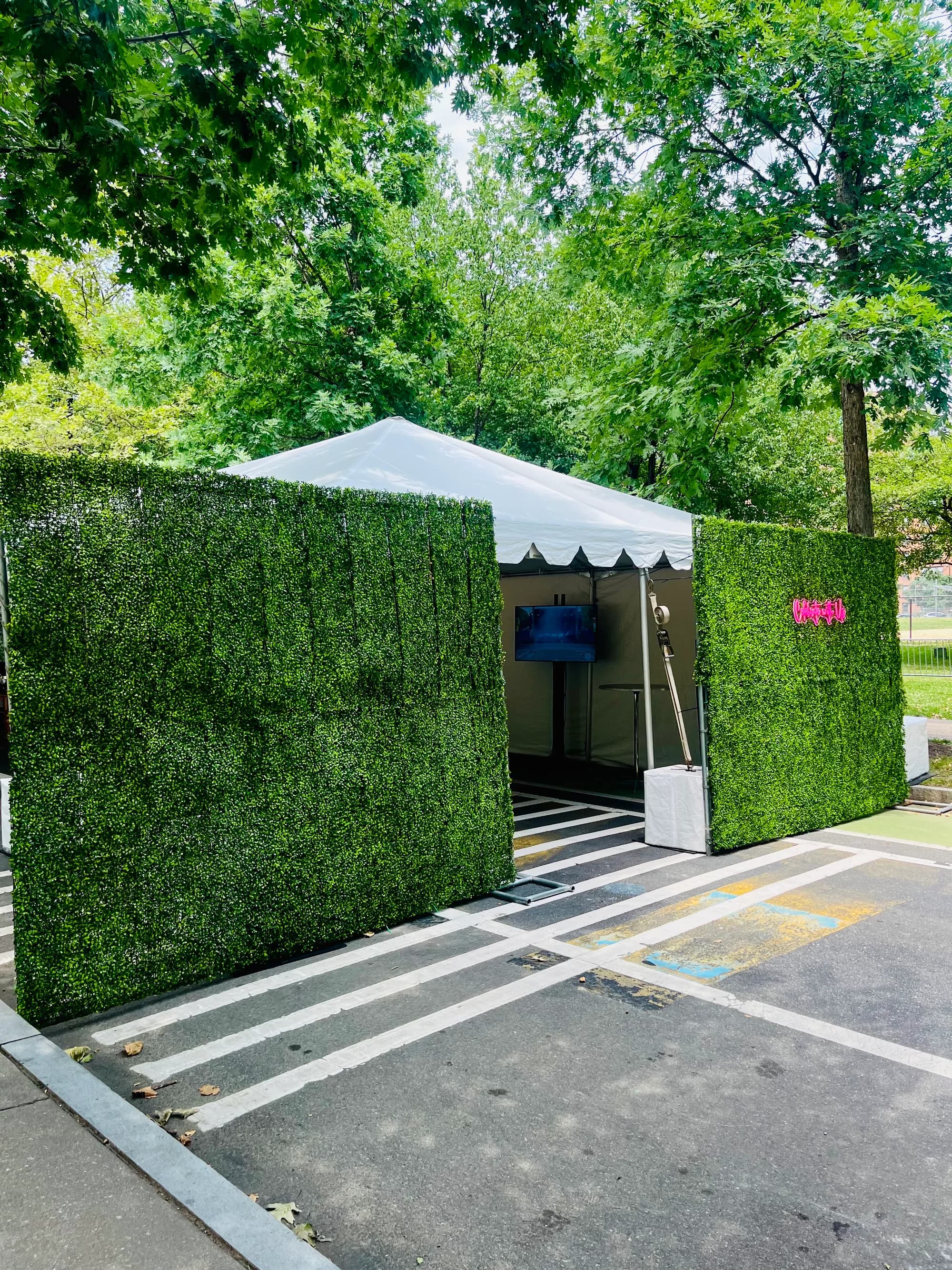 A white tent framed by two green hedge walls, a pink neon sign on one and a screen glowing inside.
