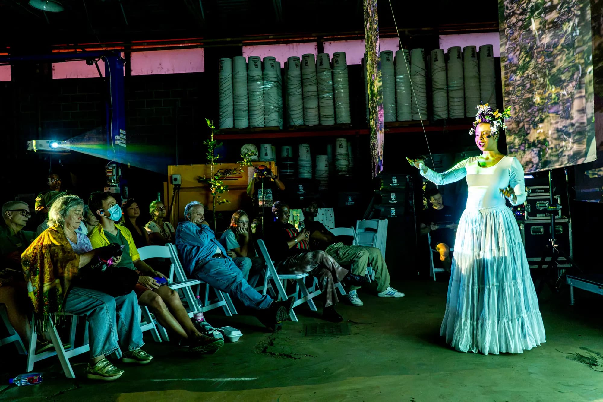 A costumed performer standing in a green-lit hall lined with tall ceramic vessels, facing a seated audience.
