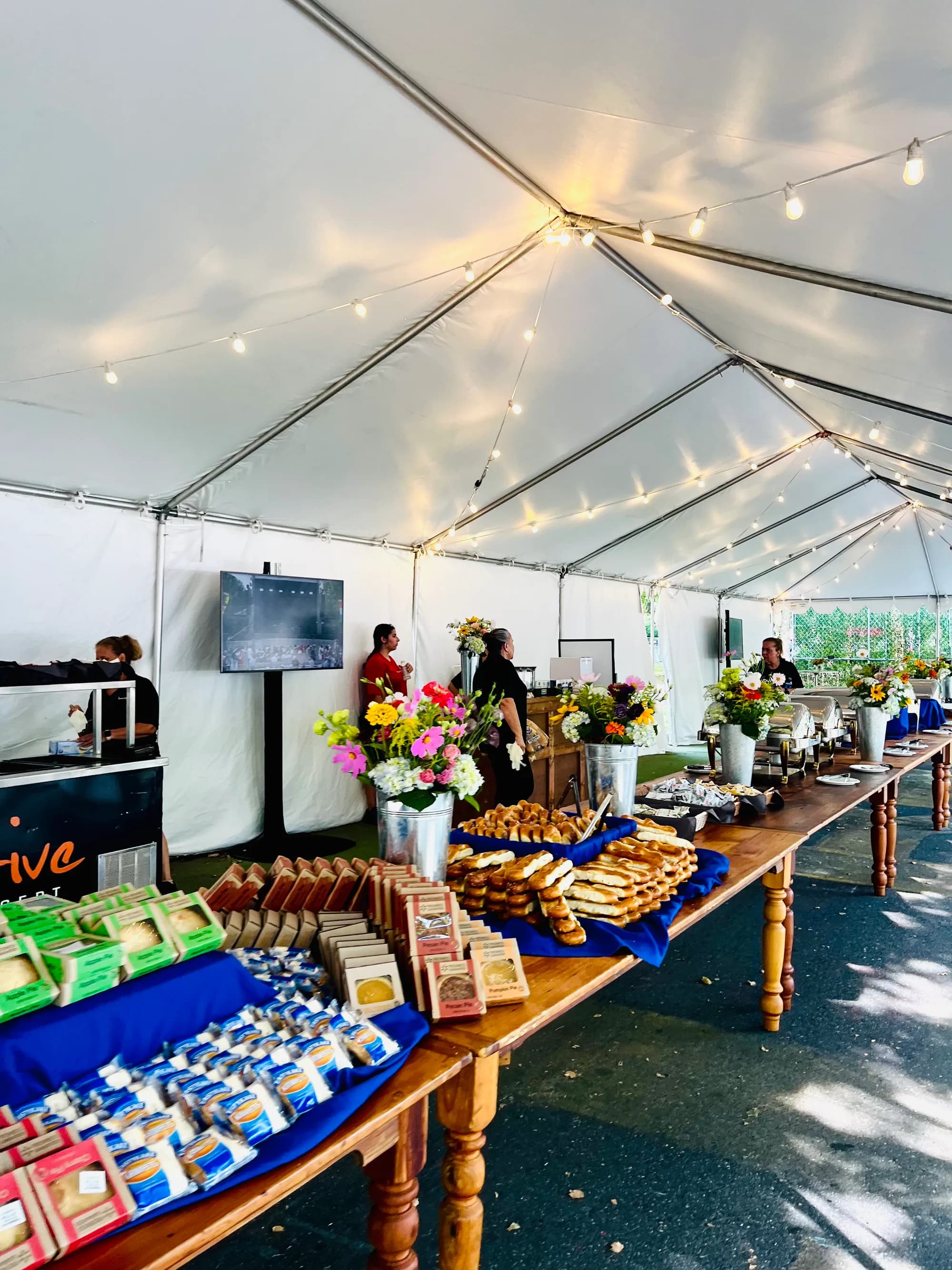 A long wooden buffet under a string-lit tent, laid with pastries, snacks, and bunches of summer flowers.