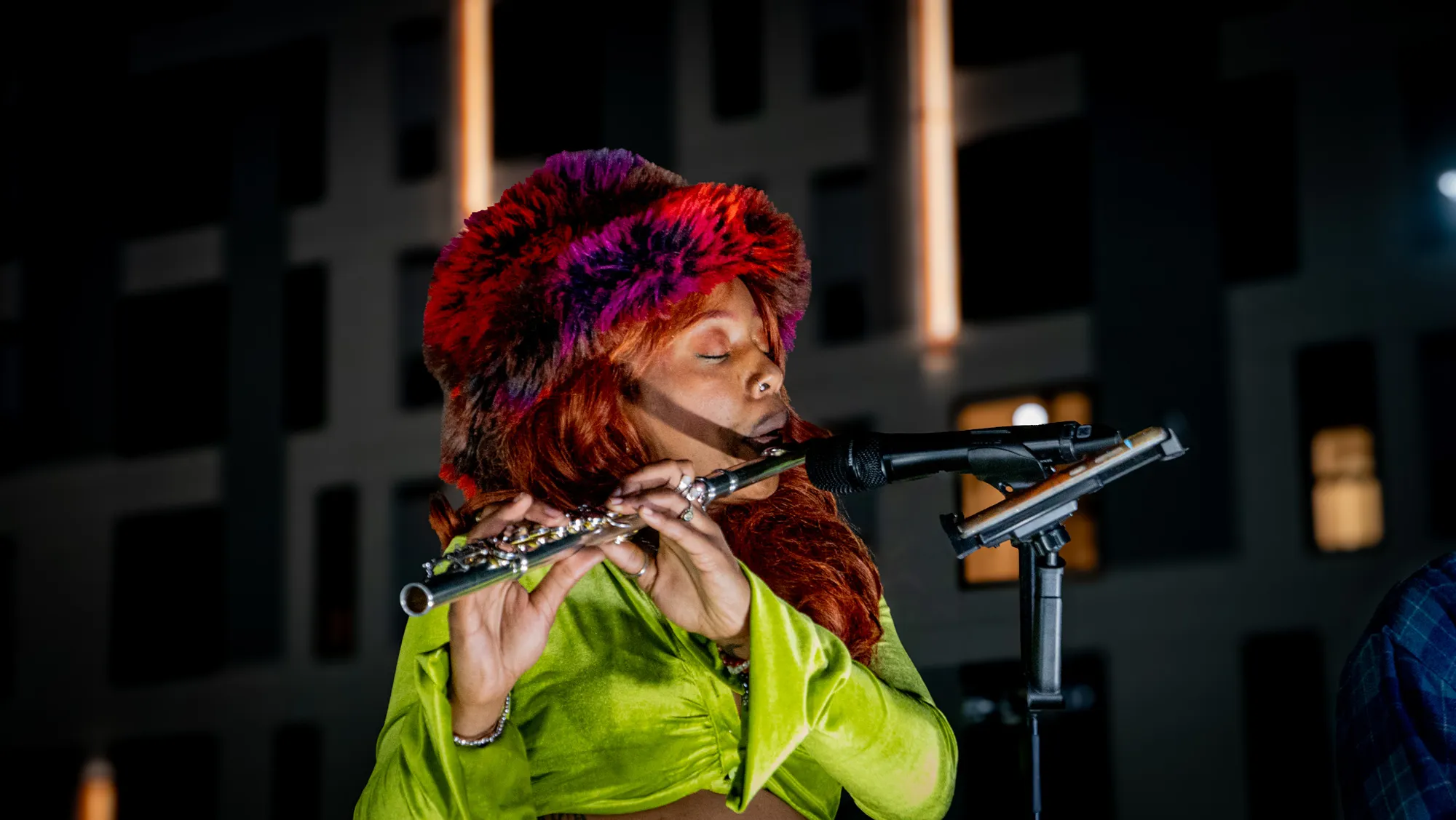 A flautist in a fur hat and lime-green top playing into the mic on the night stage.