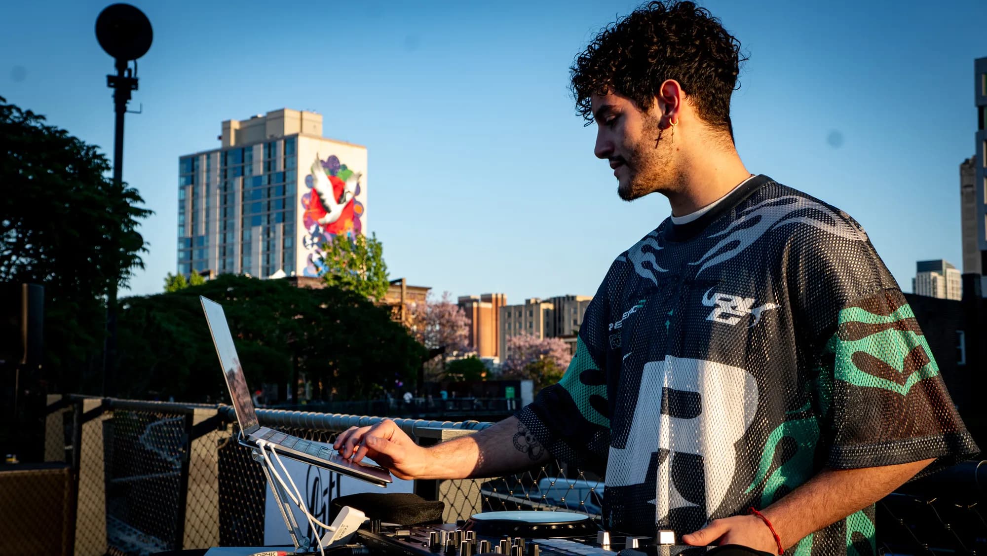A DJ at the decks at sunset, a painted high-rise mural behind him.
