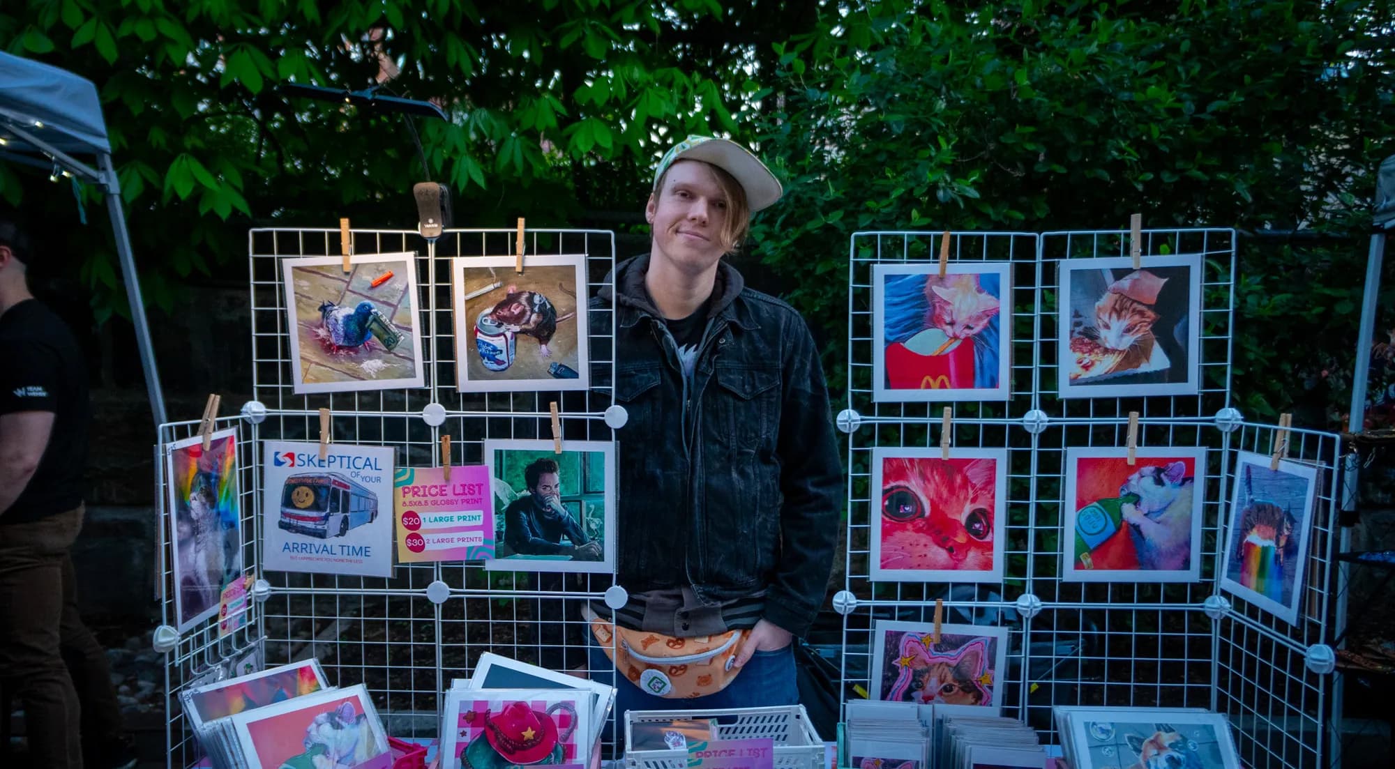 A vendor standing behind his wire grid of framed prints at the evening market.